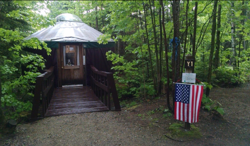 A wooden yurt entrance surrounded by greenery, with an American flag and a sign nearby.