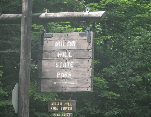 Wooden sign for Milan Hill State Park, surrounded by trees, with directions to a fire tower and picnicking area.