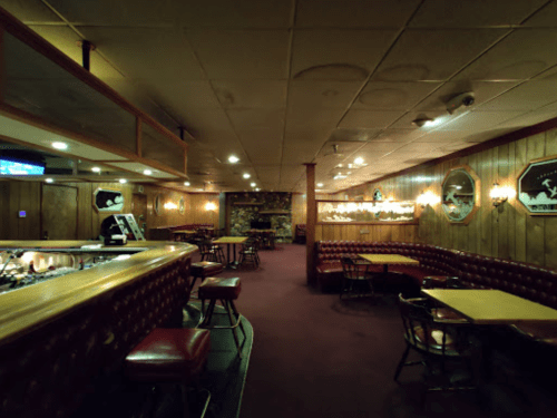 A dimly lit bar with wooden paneling, red leather seating, and tables, featuring a cozy, vintage atmosphere.