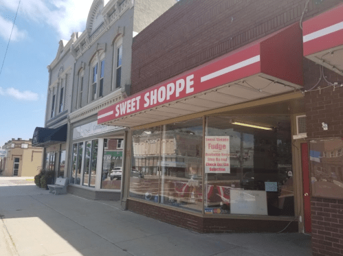 A storefront labeled "Sweet Shoppe" with large windows displaying treats, located on a sunny street.