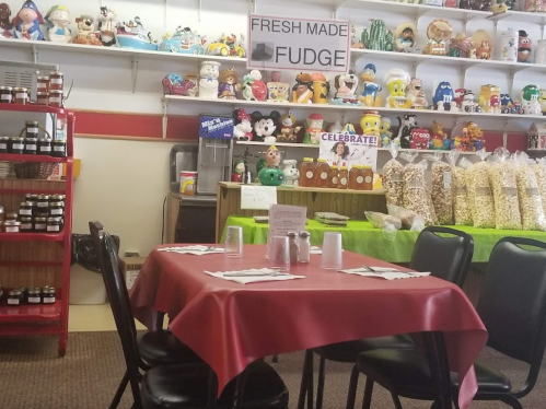 A cozy restaurant with a red tablecloth, surrounded by shelves of toys and jars, featuring a sign for fresh fudge.