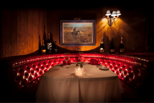 Cozy restaurant booth with red leather seating, a candlelit table, and a painting on the wall, surrounded by wine bottles.