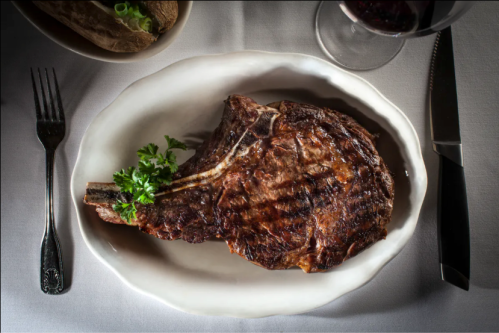 A perfectly grilled ribeye steak garnished with parsley, served on a white plate with a fork and knife beside it.