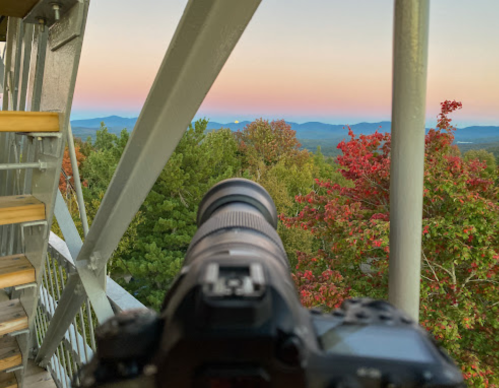 A camera on a staircase overlooks a colorful sunset with mountains and trees in the background.