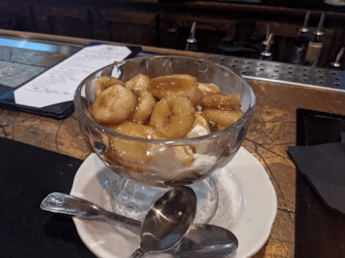 A glass dessert bowl filled with ice cream topped with caramelized bananas, served on a white plate with two spoons.