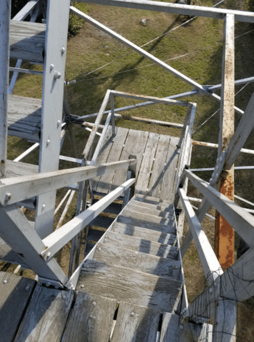 View looking down a wooden staircase on a metal tower, surrounded by grass and metal supports.