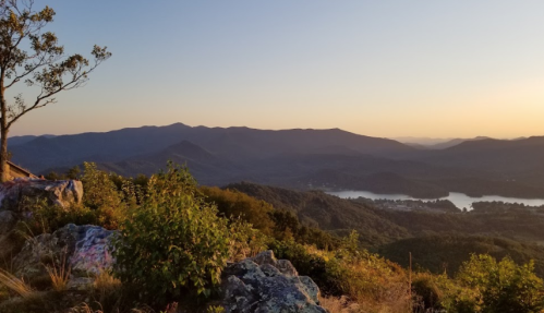 Scenic view of mountains and a lake at sunset, with trees and rocks in the foreground.