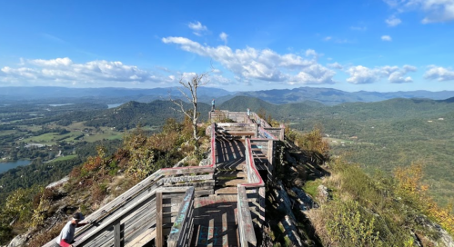 A scenic view from a mountain overlook, featuring wooden pathways and lush green valleys under a blue sky.