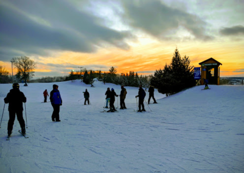 Skiers gather on a snowy slope at sunset, with trees and a ski lodge in the background.