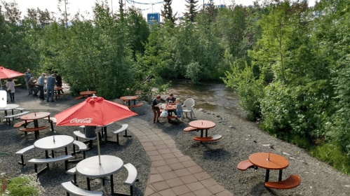 Outdoor seating area by a river, surrounded by trees, with people dining and enjoying the scenery.