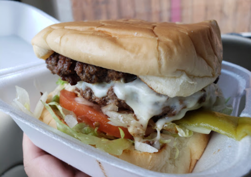 A close-up of a juicy hamburger with lettuce, tomato, mayonnaise, and pickles, served in a takeout container.