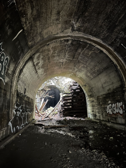 A dark tunnel with graffiti on the walls, leading to a light-filled opening and debris in the foreground.
