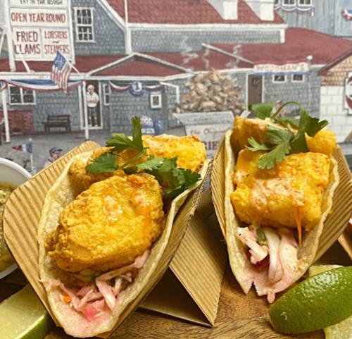 Two crispy fish tacos topped with slaw and cilantro, served with lime wedges, against a coastal restaurant backdrop.