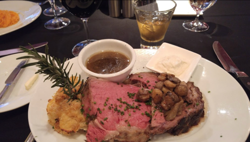 A plate featuring prime rib with mushrooms, gravy, a baked potato, and a drink, set on a black tablecloth.