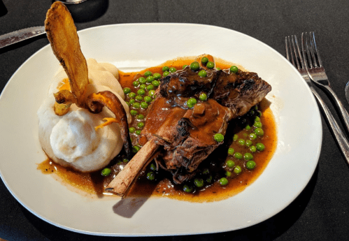A plate featuring a lamb shank with peas, mashed potatoes, and a crispy plantain garnish on a black tablecloth.