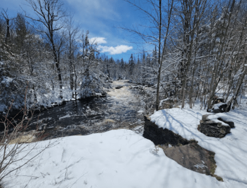 A snowy landscape with a river flowing through trees, under a bright blue sky with scattered clouds.