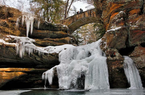 A frozen waterfall cascades over rocks, with icicles hanging and a stone bridge in the background surrounded by trees.