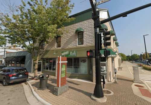 A corner building with a green sign reading "Nails," surrounded by trees and a sidewalk, near a traffic light.