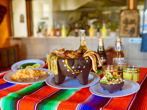 A colorful table setting featuring a traditional dish in a stone bowl, rice, salad, and drinks, with a vibrant tablecloth.