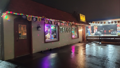 A taco restaurant at night, illuminated with colorful lights and reflecting on wet pavement.