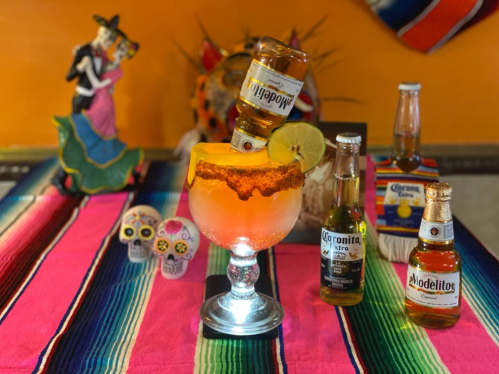 A colorful table setting featuring a large cocktail with a beer bottle, surrounded by decorative bottles and vibrant textiles.