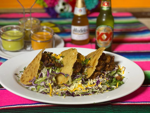 Three tacos on a bed of shredded cabbage, with sauces and two bottles of beer in the background, on a colorful tablecloth.