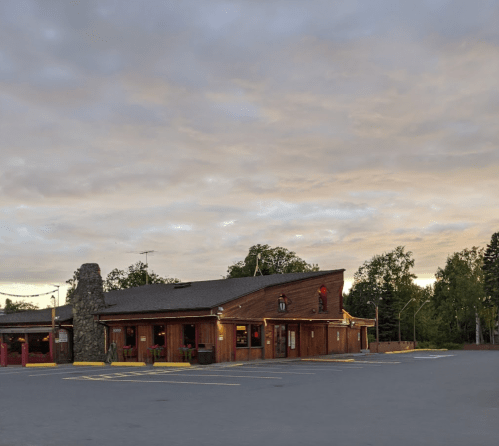 A rustic wooden building with a stone feature, set against a cloudy sky and surrounded by trees.