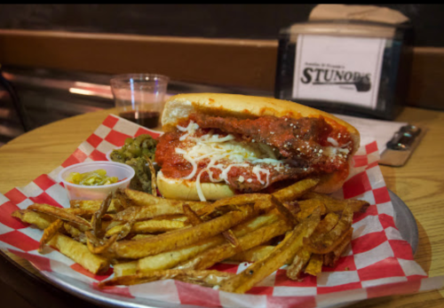 A hearty sandwich with marinara sauce and cheese, served with fries and pickles on a checkered plate.