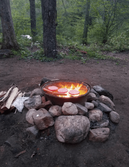 A grill over a stone fire pit with burgers cooking, surrounded by trees in a natural outdoor setting.