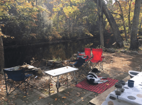 A peaceful riverside scene with colorful autumn leaves, two red chairs, a dog on a blanket, and a small table.
