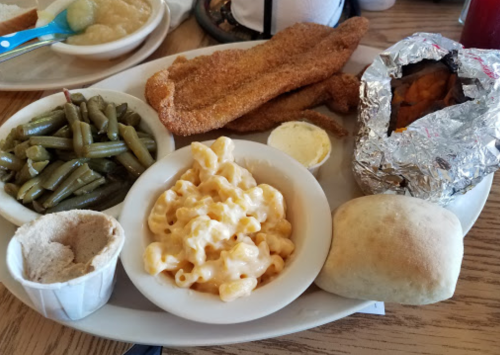 A plate with fried fish, green beans, macaroni and cheese, a baked potato wrapped in foil, and a roll.