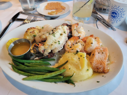 A plate of seafood featuring shrimp, lobster, salmon, green beans, and mashed potatoes, garnished with herbs.
