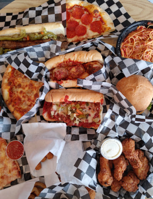 A spread of various foods including pizza, sandwiches, spaghetti, chicken wings, and dipping sauces on checkered paper.