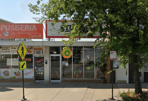 A storefront with signs for a restaurant and a delivery service, surrounded by trees and a crosswalk.