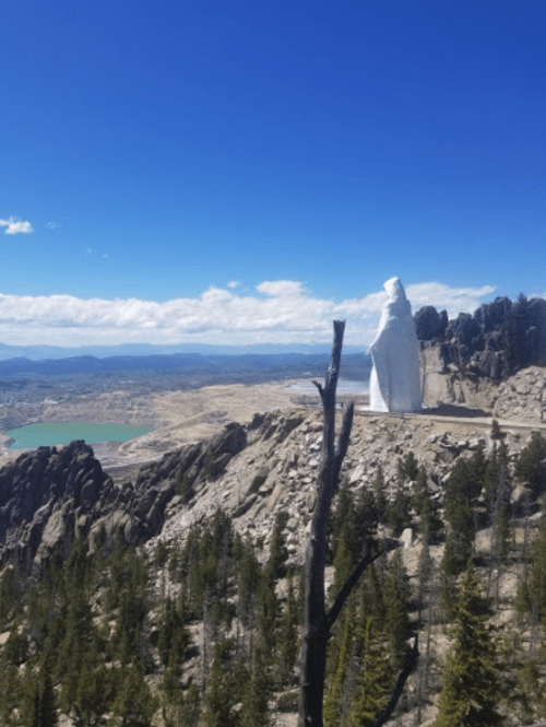 A white statue stands on a rocky outcrop overlooking a valley and lake, surrounded by trees and mountains under a blue sky.