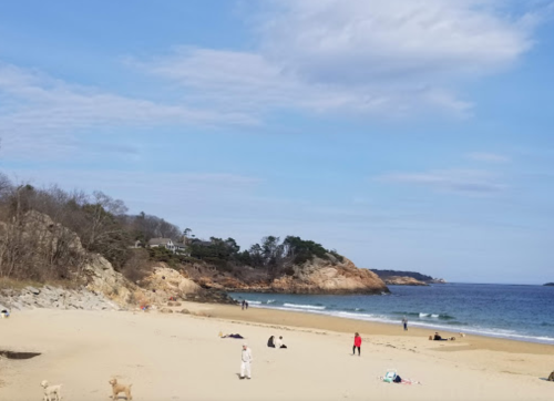 A sandy beach with people walking and relaxing, surrounded by rocky cliffs and a calm sea under a blue sky.