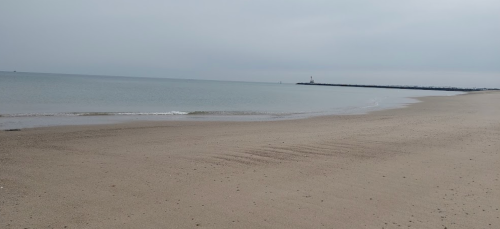 A calm beach with smooth sand and gentle waves, under a cloudy sky, leading to a distant lighthouse on the horizon.