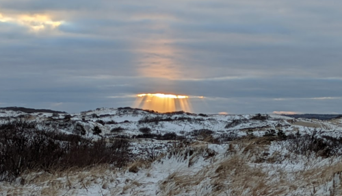 Sunset rays break through clouds over snowy hills, casting a warm glow on the winter landscape.