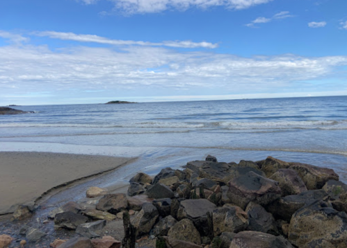 A serene beach scene with gentle waves, rocky shore, and a clear blue sky with scattered clouds.