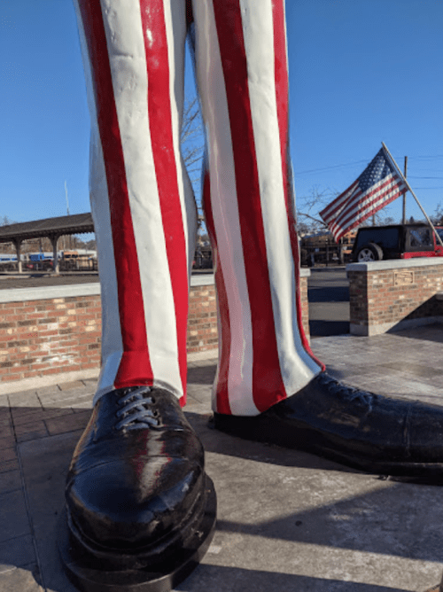 Giant statue of legs in red and white striped pants, with a large black shoe, and an American flag in the background.