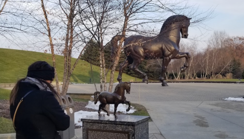 A person stands near a bronze sculpture of two horses, one large and one small, in a park setting.