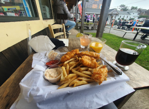 A plate of fish and fries with dipping sauces, accompanied by drinks, on a table outside a restaurant.