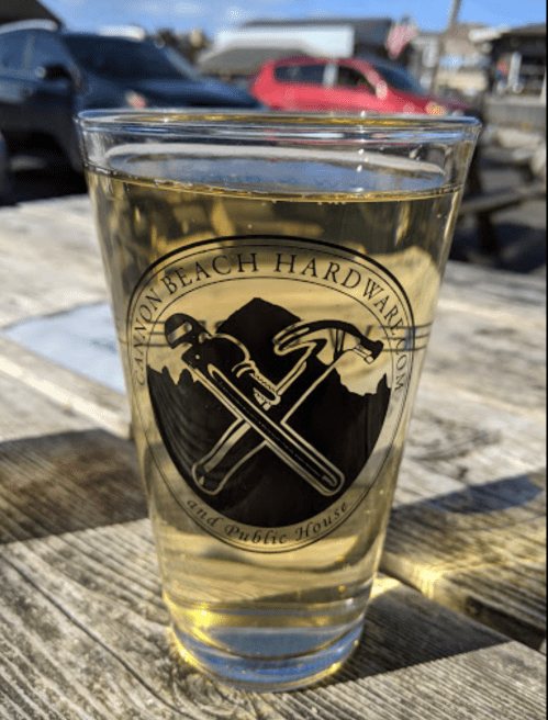 A glass of cider with a logo featuring crossed tools, sitting on a wooden table outdoors.
