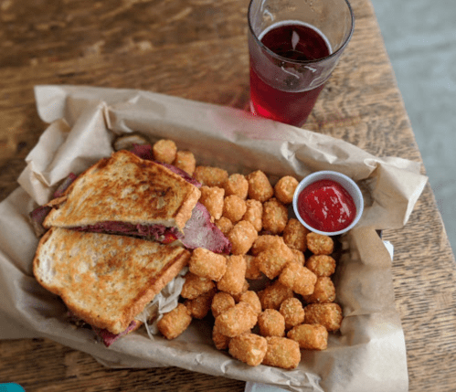 A sandwich with meat and cheese beside a pile of tater tots and a small cup of ketchup, with a drink in the background.