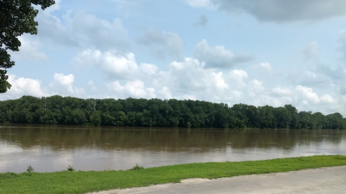 A calm river bordered by lush green trees under a partly cloudy sky.
