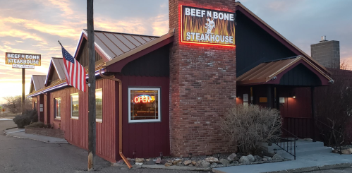 Exterior of Beef N Bone Steakhouse at sunset, featuring a sign, American flag, and warm lighting.