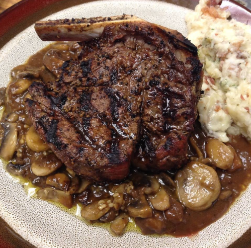 A grilled ribeye steak served with mushrooms and a side of creamy mashed potatoes on a decorative plate.