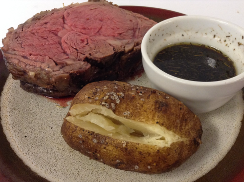 A plate with a slice of medium-rare prime rib, a baked potato, and a small bowl of dipping sauce.