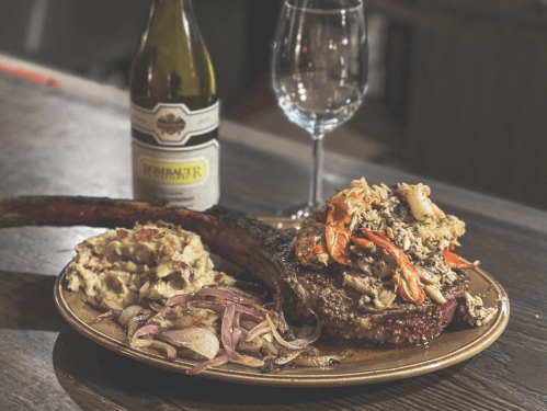 A plated meal featuring a large ribeye steak topped with seafood, sides of mashed potatoes and onions, with wine in the background.