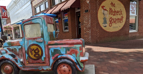 A colorful vintage truck sculpture in front of a brick building with a sign for "The Feathered Gourmet."